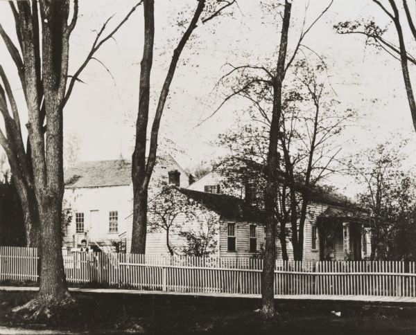 View from across street of the Whitney House. A man stands in the yard behind the fence. There are large trees along the curb, and more trees and shrubs in the yard.