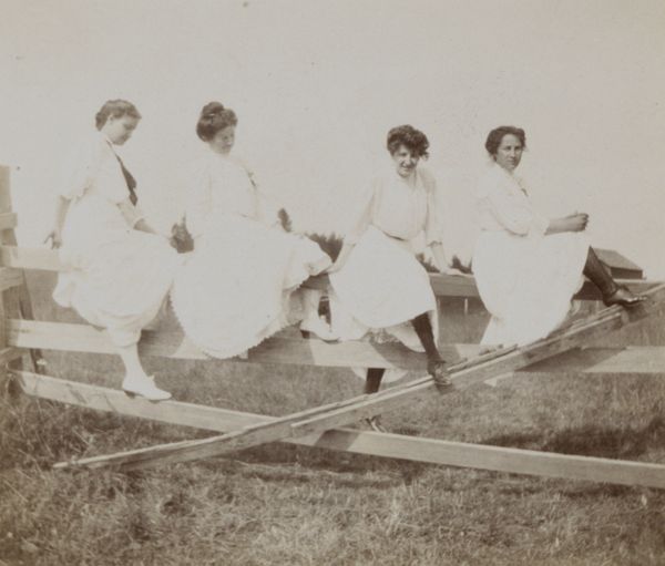 Four young women wearing long white dresses climbing and posing on a fence.