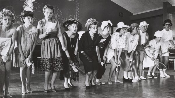 Lady members of the Brynwood Country Club performing a musical skit at the club's opening luncheon. They are concluding a historical “fashion” show, wearing various sport and “country-club wear” clothing from the 1920's to 1960.