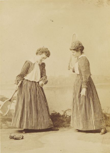 Studio portrait in front of a painted background of two young women dressed in long, striped tennis costumes and matching striped caps. They each hold a tennis racket, and the woman on the left is holding a tennis ball and pretending to set up a serve. Her hat is on the floor.