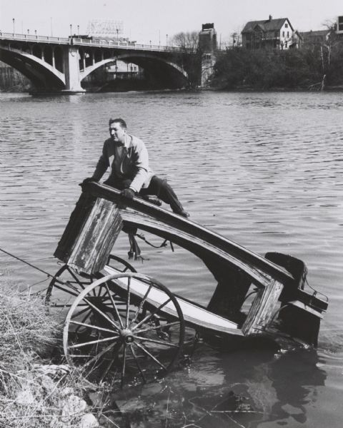 An antique 82-year-old horse-drawn hearse, pushed by pranksters into the Milwaukee River near the North Avenue bridge, being pulled out by a tow-truck, as owner Maurice Belfor rides on top for ballast. Mr. Belfor purchased the hearse from a farmer near Stevens Point.
