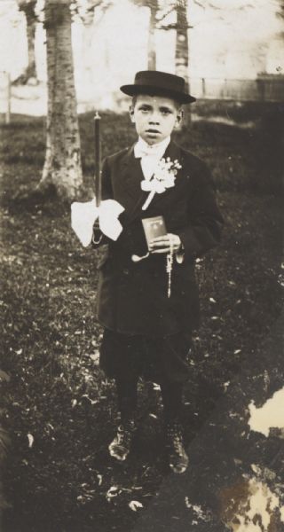 Young boy, standing in a wooded yard, dressed for his first communion with a boutonniere pinned to his lapel. He is holding a candle, rosary and prayer-book.