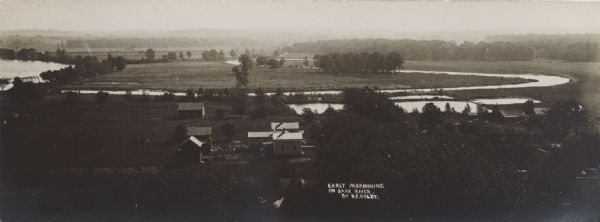 Elevated view over the ox-bow of the Bark River in early morning. Buildings, dwellings and barns are in the foreground at the bottom of the hill, and a bridge is on the far left.