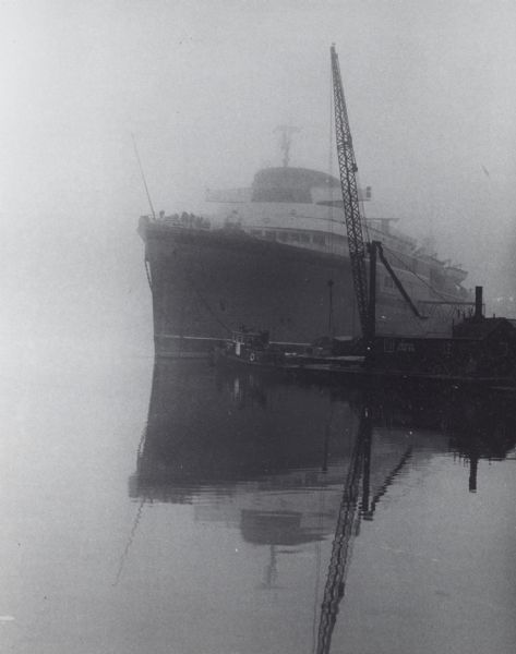 Lake steamer at dock, in fog. To the right is the boom of a crane. In front of the steamer is a tugboat.