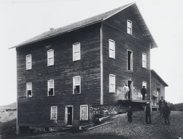 Exterior view of a mill owned by George Mills Hull. Three men stand in the foreground, and three more stand near the entrance to the mill.