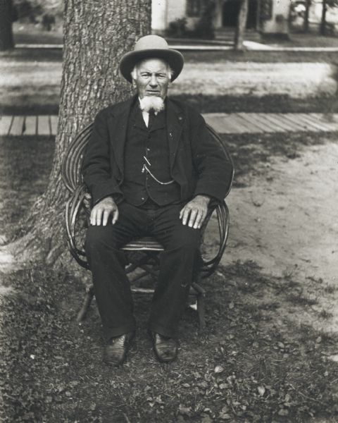 Daniel Reynolds, wearing a hat, suit coat, and vest with watch fob, sits in a bentwood chair in front of a large tree.  There is a board sidewalk behind him and a house across the street.