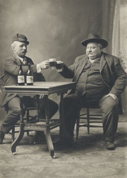 Two men, posed, seated at a table toasting each other with a mug of beer.