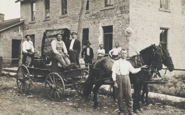 A group of unidentified men standing with a horse and wagon. The wagon is carrying a barrel of beer. The sign on the building in the background is an advertisement for beer.