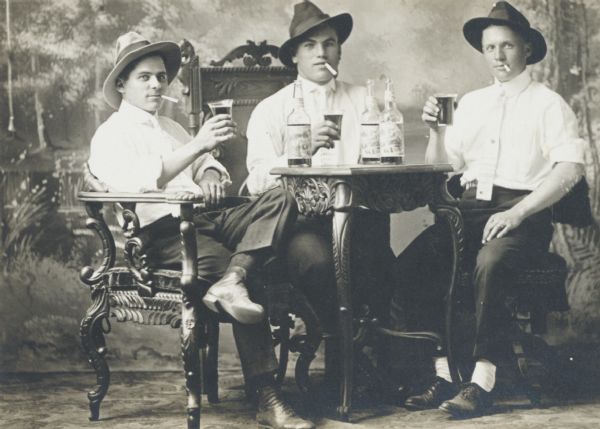 Studio triple portrait in front of a painted backdrop: young men with hats askew, drinking beer and smoking.