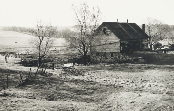 View down hill of a farmyard. A pasture with several trees is visible, in addition to two farm buildings.