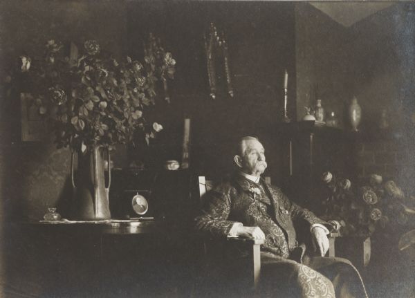 Unidentified elderly man wearing smoking robe, seated at rest in half light in an elaborate residence.