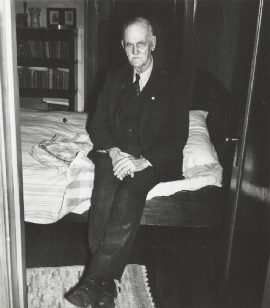 Unidentified elderly man seated on bed.