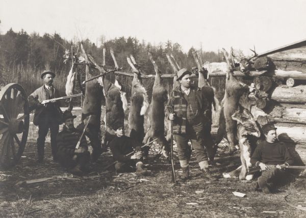 Five men holding hunting rifles sitting or standing to the left of a log cabin (only a corner of which is in the image) in front of the hanging carcasses of seven or eight deer, as well as the carcasses of some birds and smaller mammals such as rabbits. Patches of snow are on the ground. In the background is a dense stand of pine and leafless deciduous trees.