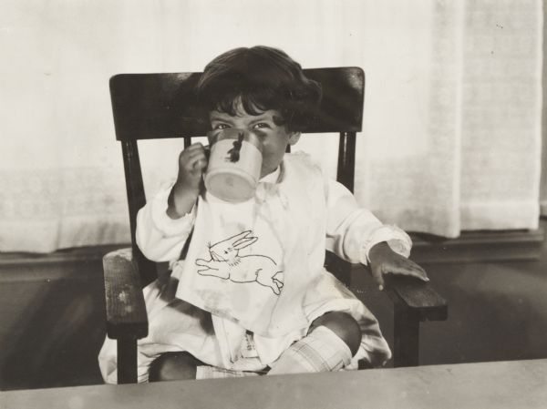 Child sitting in a high chair drinking tomato juice from a mug. The child is wearing a bib with a rabbit stitched on the front.