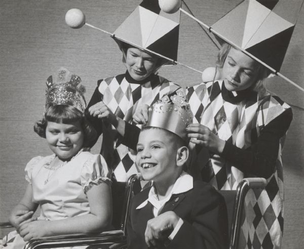 Costumed figures placing crowns on the heads of two children in wheelchairs, probably as king and queen at an institutional party.