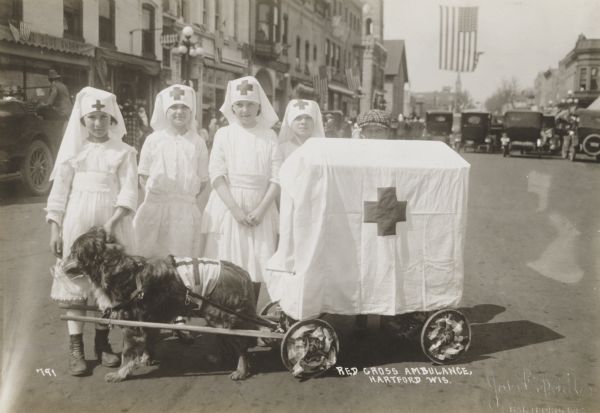 Young girls costumed as Red Cross nurses for a parade, with a small wagon drawn by a pet dog decorated as a field “ambulance.”