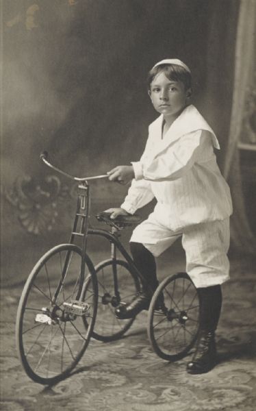Full-length studio portrait in front of a painted backdrop of a young boy with a tricycle.