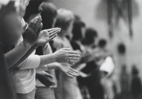 “Close-up” photograph of a line of people, a detail showing essentially their hands clapping.
