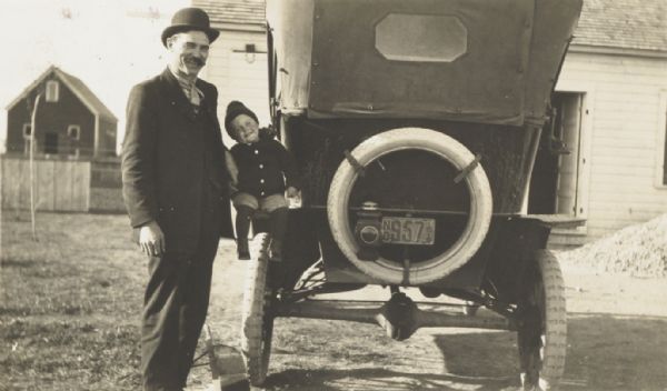 Man and small boy posing at the back of a Ford automobile in a farm yard.