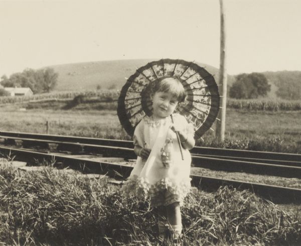 Unidentified small girl holding a parasol, posed beside railroad tracks.