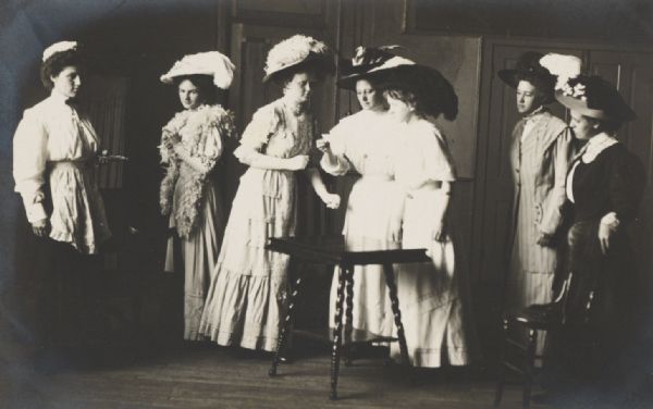 Seven women on stage, apparently in a scene from an amateur theatrical performance, wearing conventional street clothes.