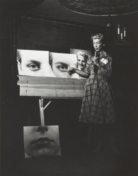Woman on stage with microphone and visual props, apparently demonstrating eye and other makeup techniques.