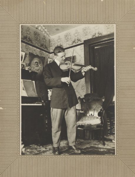 Young man standing in a turn-of-the-century parlor interior playing a violin.
