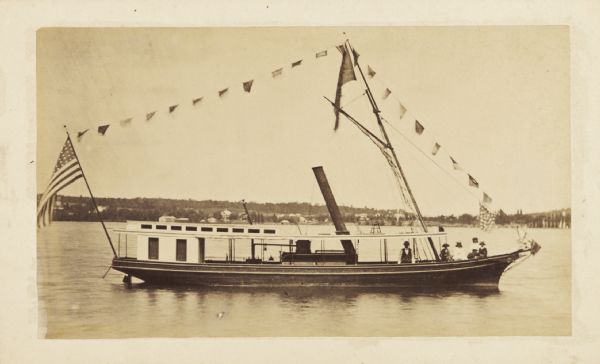 Small steam yacht decorated with pennants on Lake Geneva. A small group is at the front of the yacht. Shoreline with boathouses and dwellings can be seen in the far background.