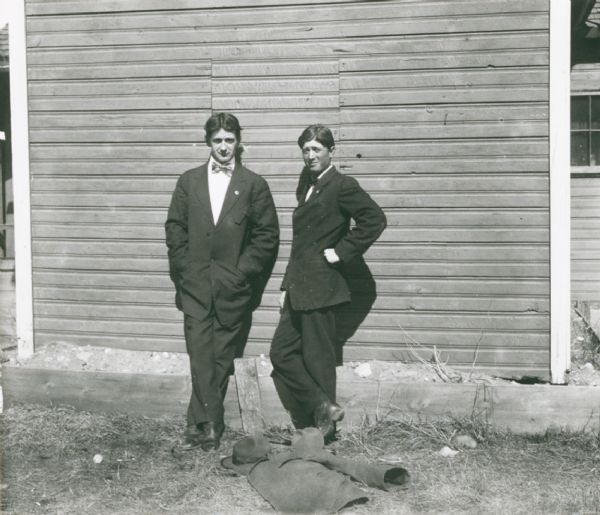 Two young men in a cynical pose, leaning against a siding wall, with a mock grave (headboard, hat and coat) in the foreground.