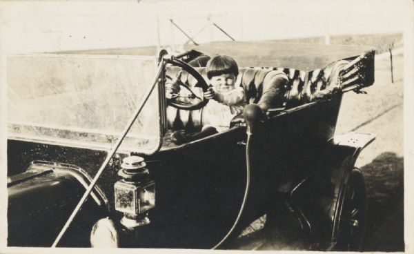 Child sitting in a Model T Ford touring car, grasping the steering wheel.