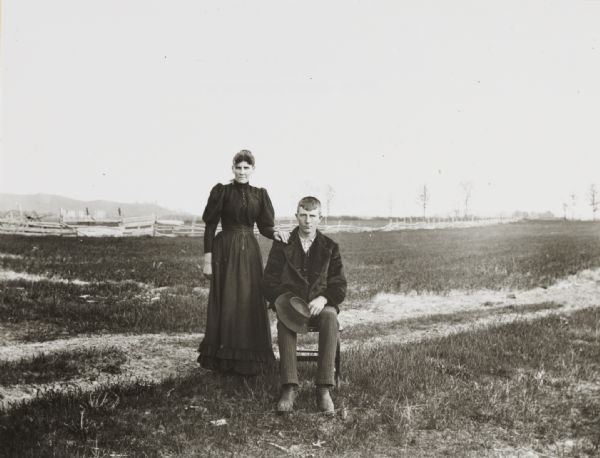 Posed portrait of a young couple outdoors in a field. The man is seated, the woman is standing beside him, with a wooden fence in the background.