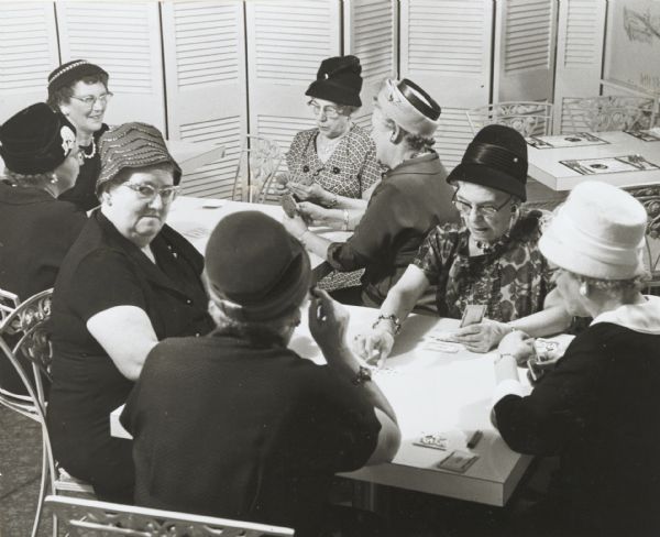 Group of women playing cards while wearing hats and pearls.