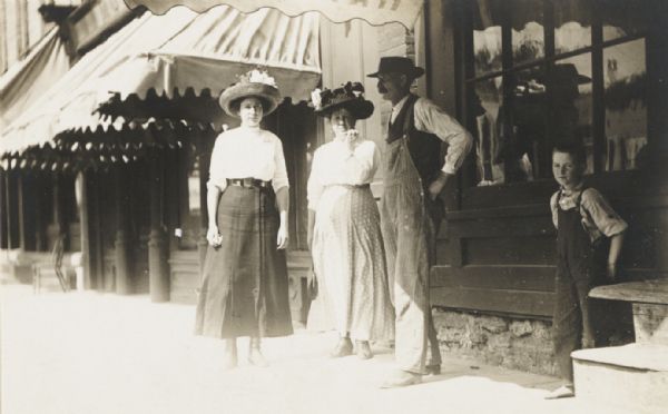 Two women, a man and a boy standing informally in front of a row of downtown stores in a small town.