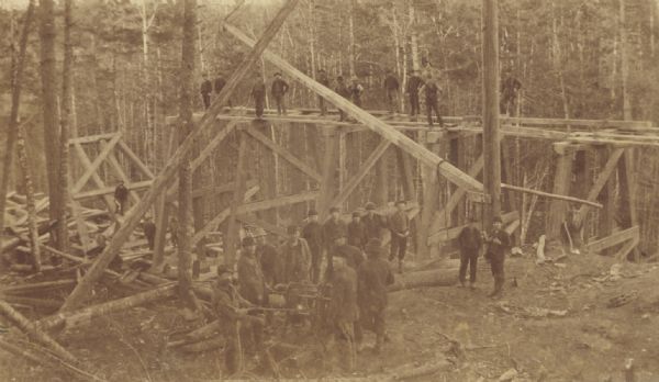 Bridge carpenters and other workmen with a winch constructing a railroad trestle over a wooded valley.