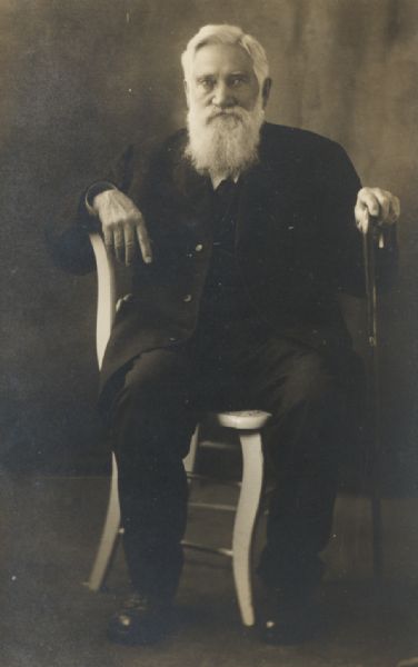 Studio portrait of Asa Curtis Bennett, sitting in a chair and holding a cane in his left hand. He was a pioneer in the cranberry growing industry in Wisconsin.
