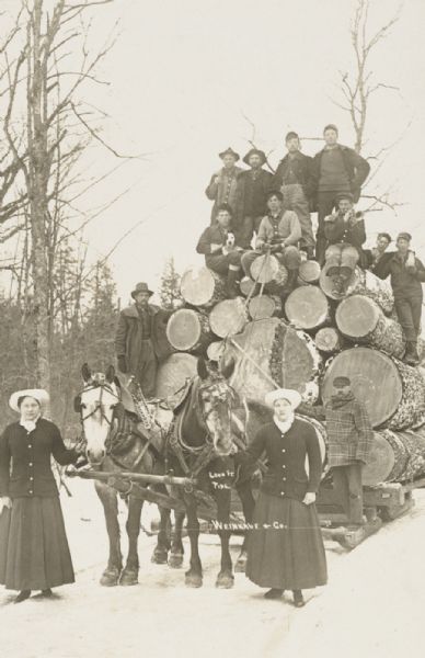 Horse-drawn load of 6000 ft. of pine logs, accompanied by a cornet player and two women, at the Weinkauf & Co. timber lands.
