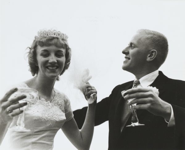 Young couple drinking champagne, perhaps at their wedding. They are both holding champagne flutes, and the woman is wearing a wedding dress and a veil, and holding gloves in her left hand.