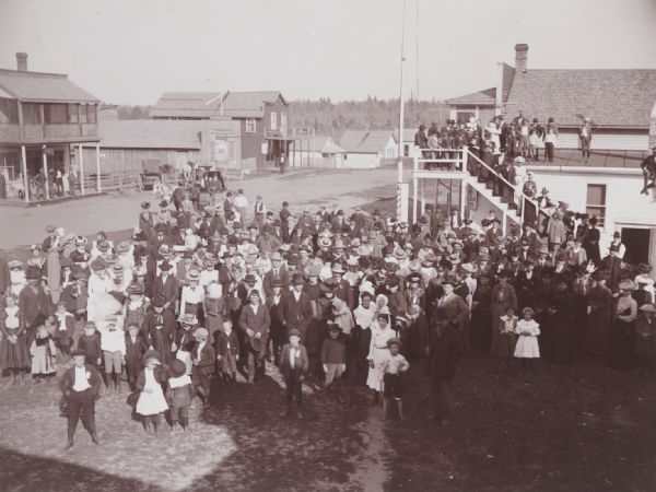 Elevated view of crowd of men, women and children, gathered for some unidentified occasion, standing in the street. There is also a group of people gathered on the exterior steps and roof of the building across the street.