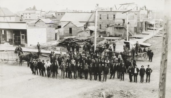 Elevated view of group gathered and posed on a new street. Behind the group is a large pile of lumber, wooden sidewalks, and what appear to be newly-constructed buildings. More buildings are in the background.