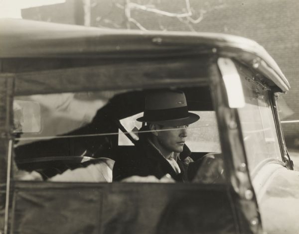 View through passenger side window of a young man seated at the steering wheel of an automobile involved in a shooting case.
