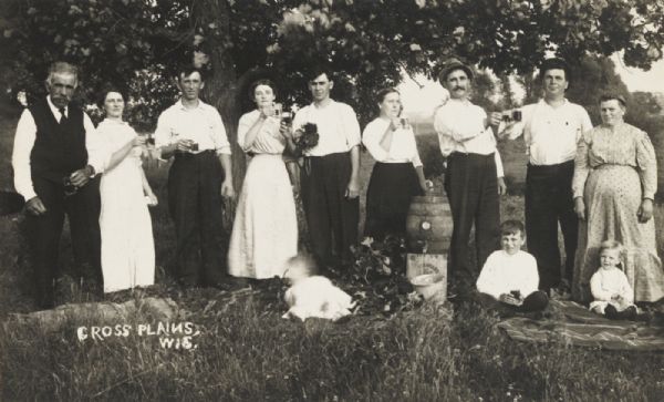 Posed line of alternating men and women in an orchard standing drinking cider of beer drawn from a keg in the center. Three young children are sitting on the grass in the foreground.
