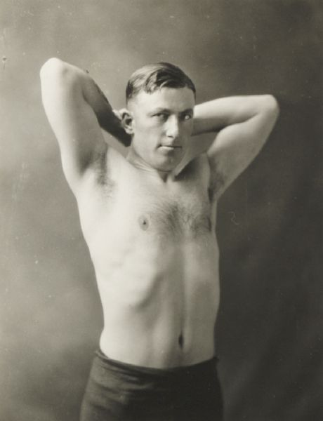 Young man, bare to the waist, with his hands behind his head, posed to show his muscles.