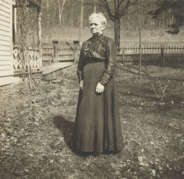 Elderly woman posed standing in the yard of a frame residence.