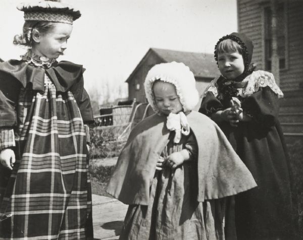 Everetta Bass (dressed in plaid), standing with two girls wearing capes and bonnets. The smallest of the girls is identified as Verta. There is a baby carriage behind them. They are standing on a board sidewalk, and there is a house and other buildings in the background.