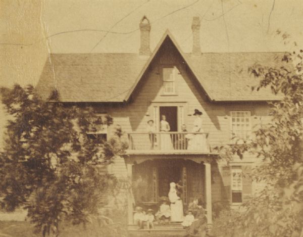 Shingled frame residence of A.G. Ellis. Children and adults are posed on the front porch, and on a balcony above.