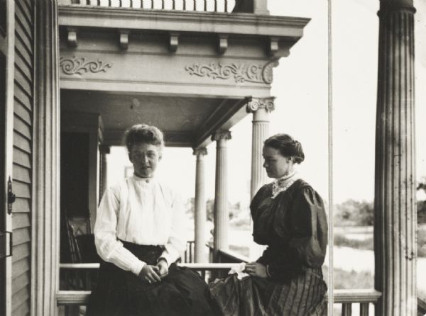Mary E. Smith, right, and a friend seated on the railing of a neo-classical residential front porch. There is another porch in the background.
