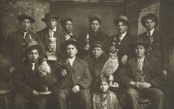 Posed studio group portrait in front of a painted backdrop of Winnebago Indian men and one young woman. Each of the men is holding a mounted bird of some species.  