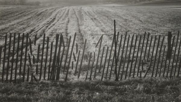 A wooden snow fence lines a large field. Trees and hills are in the background.