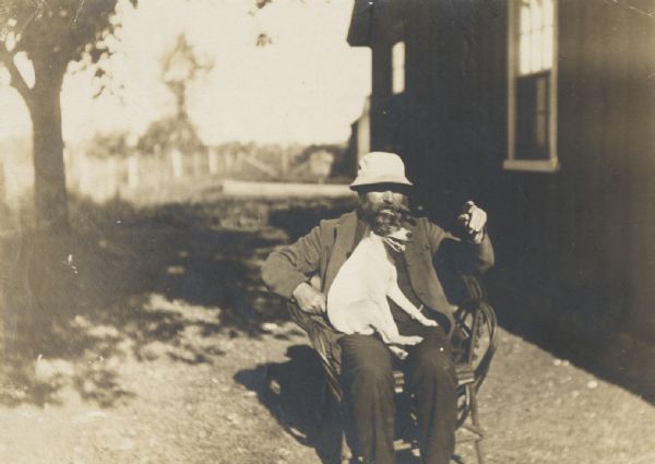 Informal portrait of “Mr. Spangenberg, RR bridge tender”, sitting in the sun with his dog on his lap, talking and pointing. He is wearing a hat and has a pipe in his mouth.
