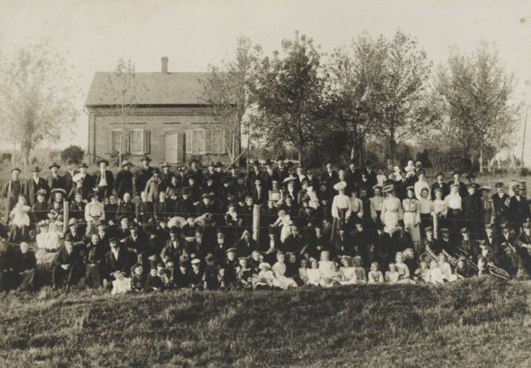 Large group of men, women and children, unidentified, posed with members of a brass band at a gathering. The group is posed in front of and behind a fence, and there is a brick building in the background.